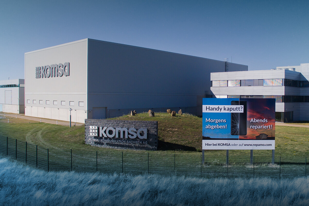 Firmenzentrale der KOMSA mit dem Logo an der Fassade und Werbetafeln auf der Wiese davor