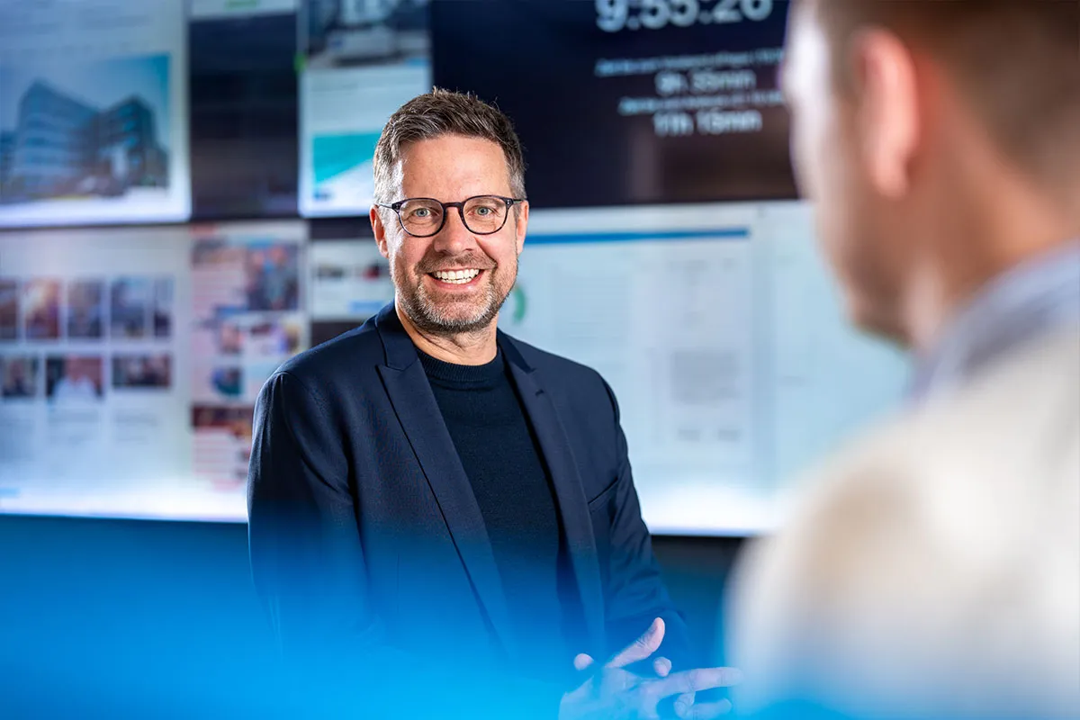 Mitarbeiter im Sakko lacht im Gespräch vor der digitalen Medienwand der Freien Presse