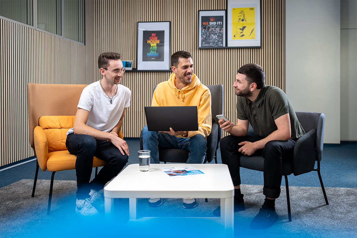Drei Kollegen im Gespräch in der Sitzecke, einer mit Notebook auf dem Schoß