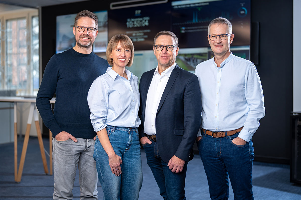 Gruppenaufnahme zu viert vor der Monitorwand des Newsrooms