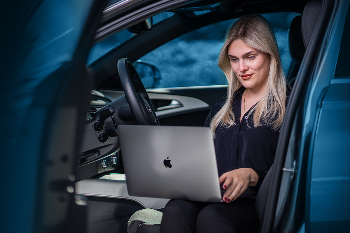 Mitarbeiterin arbeitet am Notebook auf dem Beifahrersitz, im Hintergrund das Firmengelände