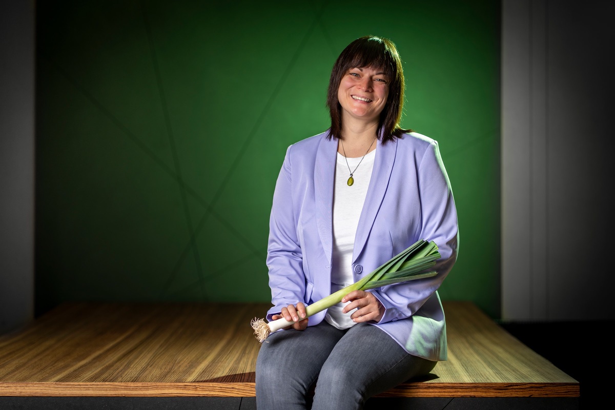 Anja Lauchart im lila Blazer sitzt auf einem Holzpodest vor grüner Wand und hält eine Lauchstange im Arm.