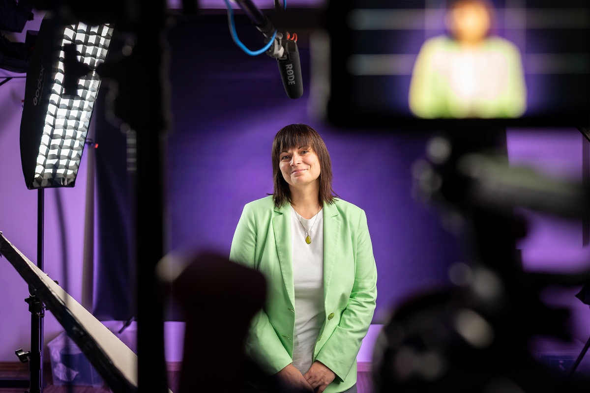 Anja Lauchart steht im hellgrünen Blazer vor lila Hintergrund, im Vordergrund unscharf Leuchte, Kamera und Monitor.
