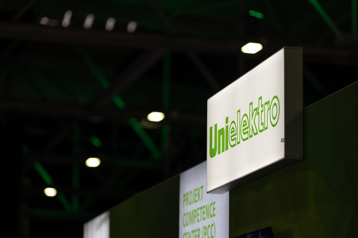 Unbearbeitete Aufnahme: Leuchtkasten mit dem UNI ELEKTRO Logo am Messestand, die Hallendecke dahinter fast schwarz