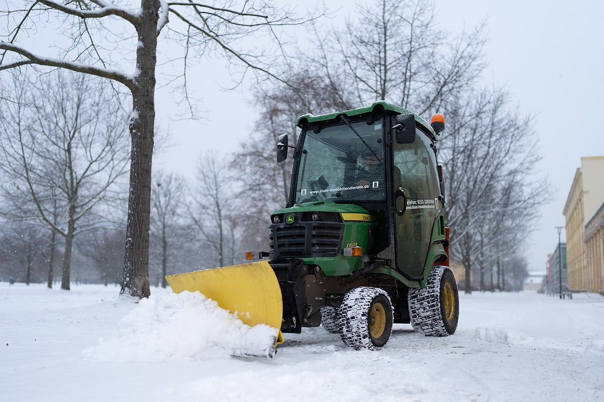 Unbearbeitete Aufnahme: Kleintraktor von Blum Immobiliendienste mit gelbem Räumschild schiebt Schnee auf einem Parkweg