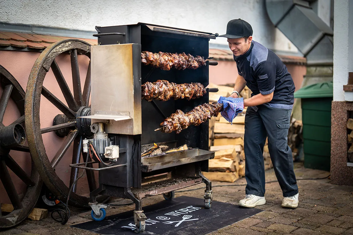 Mitarbeiter bereitet Spießbraten am Grill vor dem Hotel zu