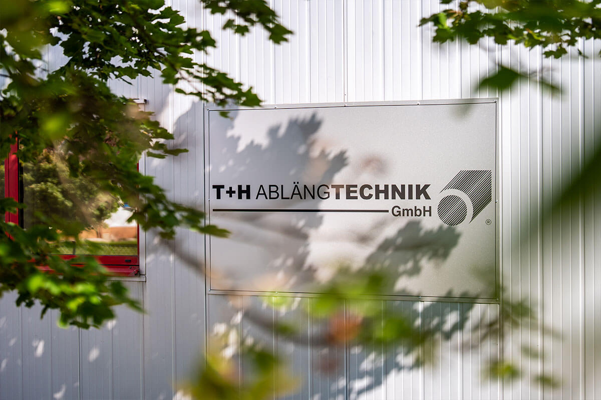 Firmenschild der T+H Ablängtechnik an der Hallenwand, durch Laub fotografiert